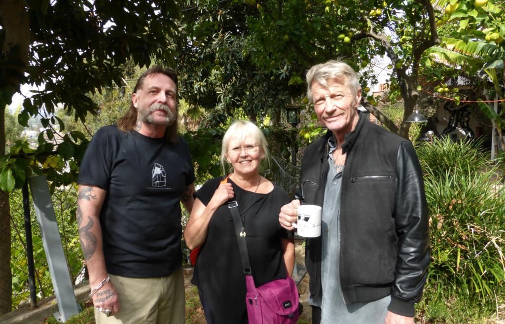 Kuvassa vasemmalla S.R. Sharp, jolla on viikset ja pitkä tukka. Hänellä on musta t-paita. Minä seison hänen vieressään ja minullakin on musta t-paita ja kameralaukku kaulassa. Oikealla seisoo Durk Dehner, jolla on viikeset ja harmaa tukka. Hänellä on kädessä kahvimuki ja päällä musta nahkatakki ja sen alla harmaa paita. Seisomme auringon paisteessa vihreässä puutarhassa. Taustalla on appelsiinipuu.