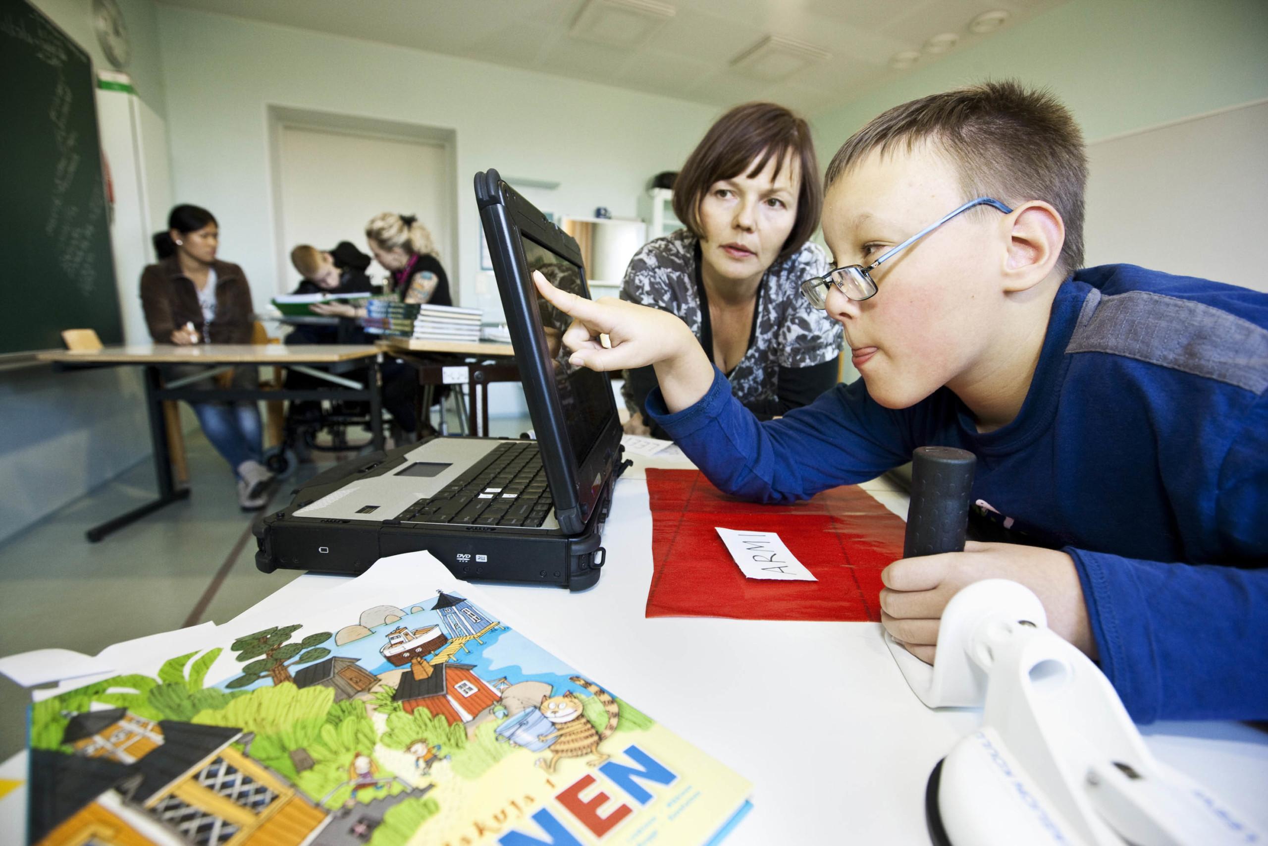 Poika tutkii etusormella kosketusnäyttöä koulussa. Taustalla opettaja tarkkailee hänen työskentelyään ja katsoo myös näyttöä.