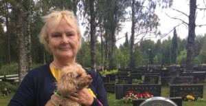 Lyhyttukkainen vaalea, keski-ikäinen nainen seisoo hautausmaalla sylissään pieni vaaleanruskea villakoira