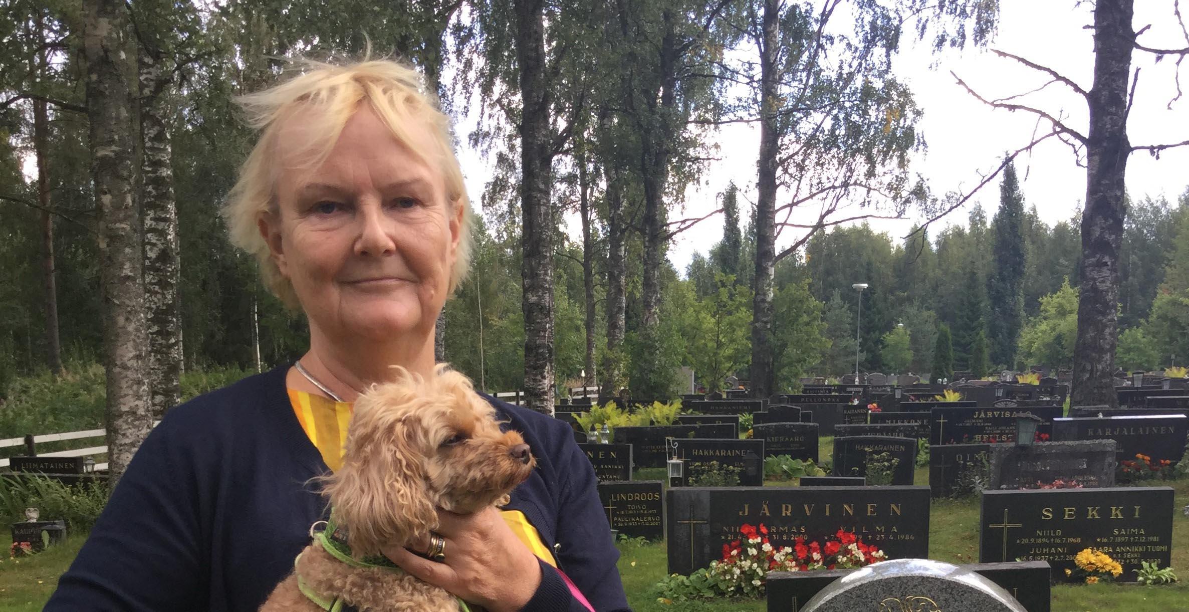 Lyhyttukkainen vaalea, keski-ikäinen nainen seisoo hautausmaalla sylissään pieni vaaleanruskea villakoira