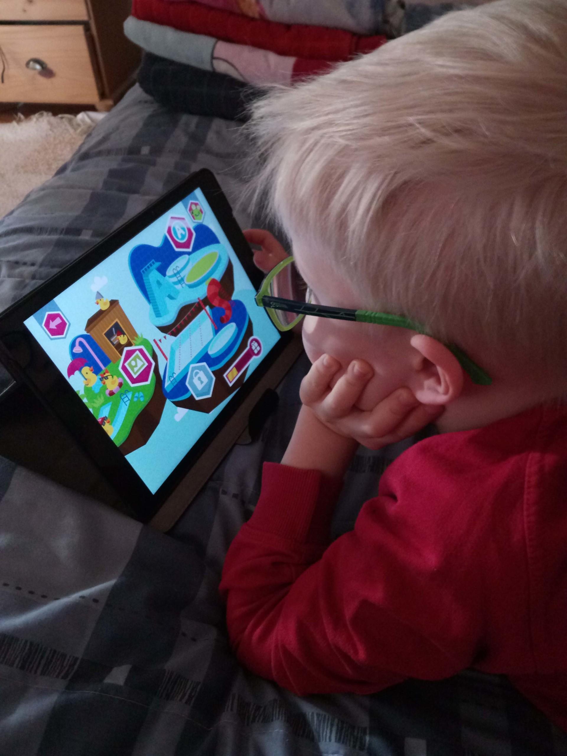 Kuvassa punapaitainen poika pelaa taböetilla värikästä oppimispeliä. Pojalla on vaaleat hiukset ja vihreät silmälasit. Poika on selin kameraan.