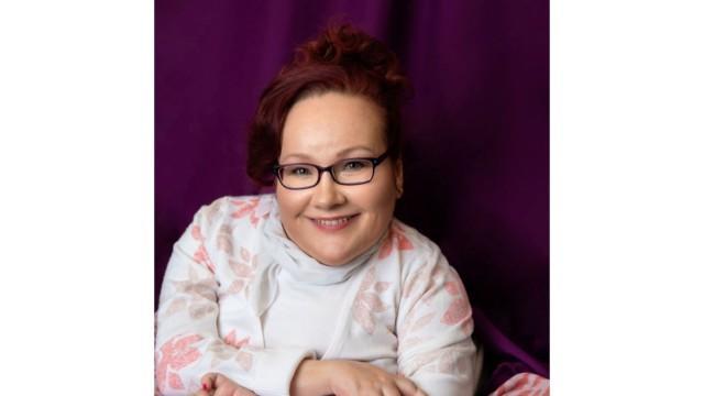 Kuvassa hymyilevä naishenkilö, jolla on punainen tukka, vaalean sävyinen paita sekä tummat silmälasit