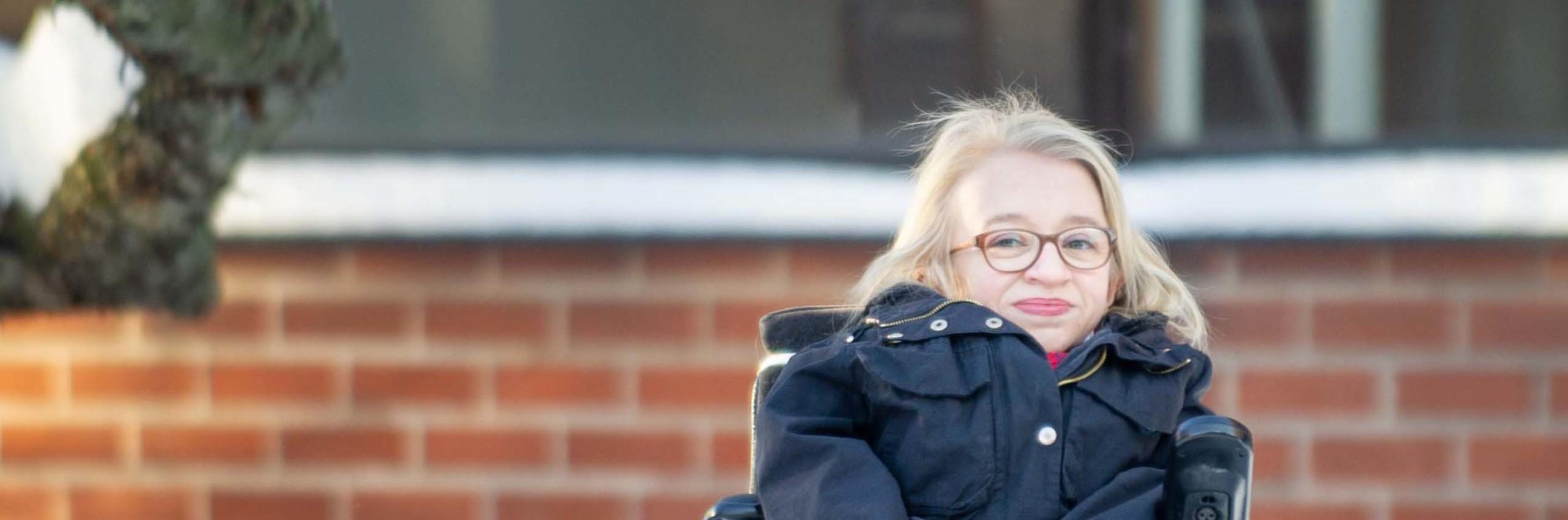 Vaalea- ja pitkätukkainen, silmälasipäinen nainen istuu tummassa takissa pyörätuolissa. koulun pihalla.