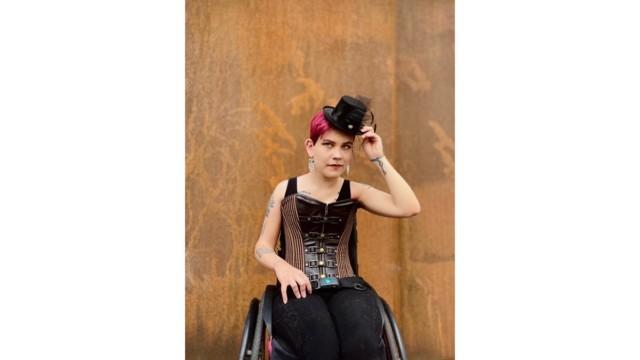 Mustiin pukeutunut, pyörätuolissa istuva henkilö nostaa päästään musta hattua